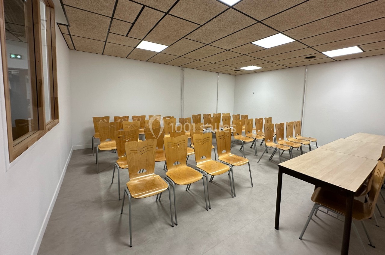 Salle de réunion avec des chaises en bois alignées face à une table, éclairée par des plafonniers.