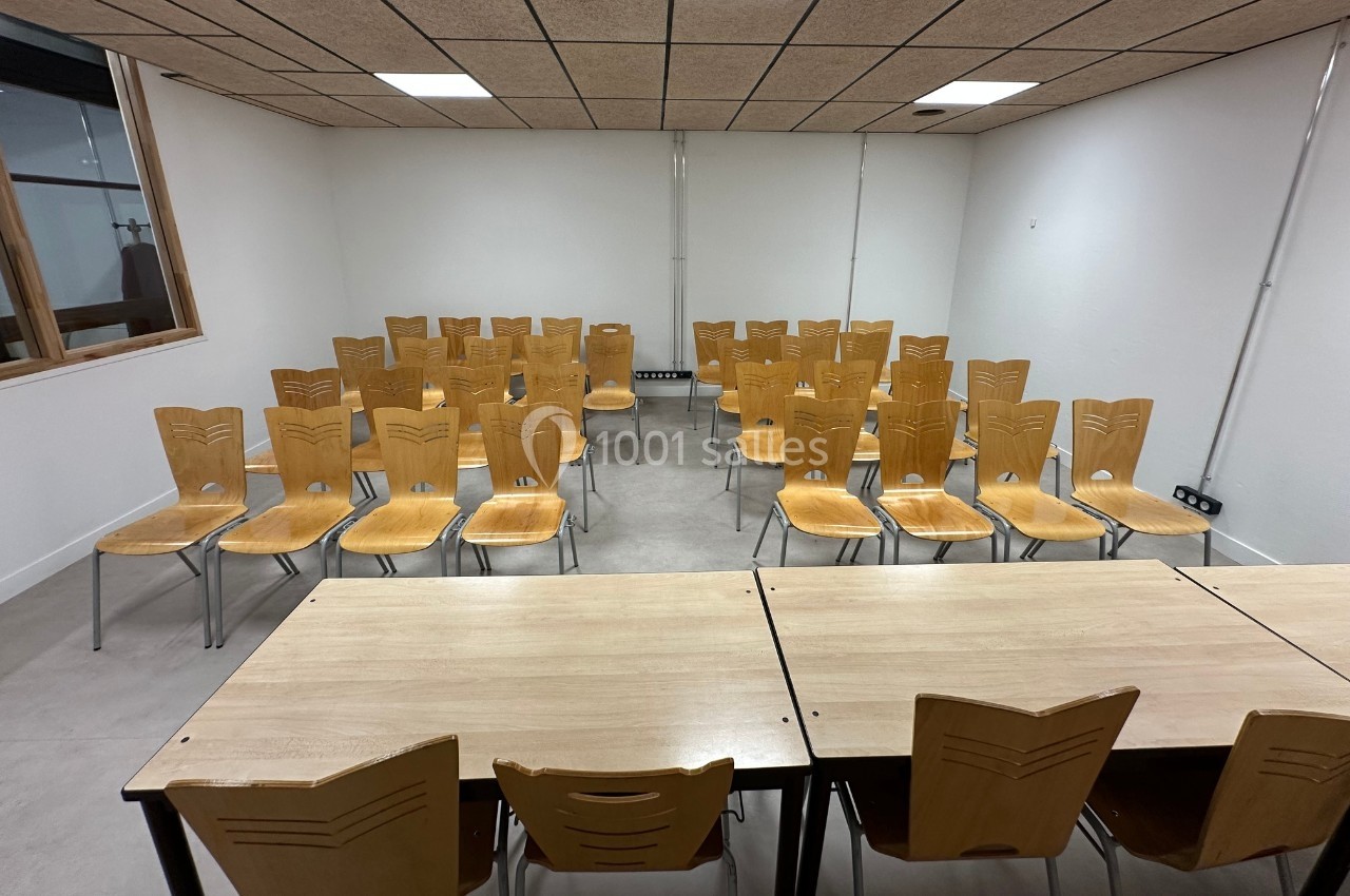 Salle de réunion avec des chaises en bois disposées en rangées face à des tables, murs blancs et plafond en dalles.