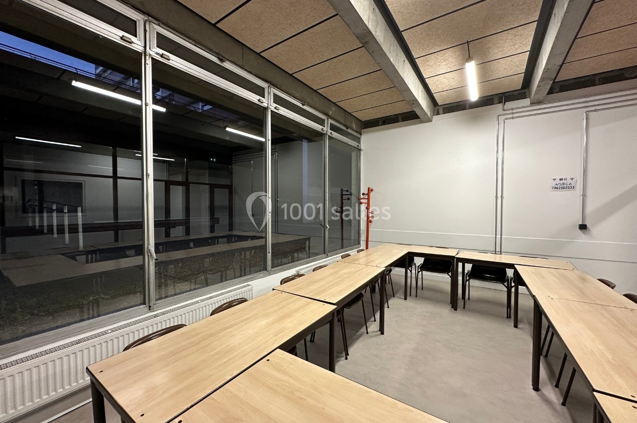 Salle de classe vide avec des tables disposées en U, éclairée par des néons et une grande baie vitrée donnant sur un couloir.