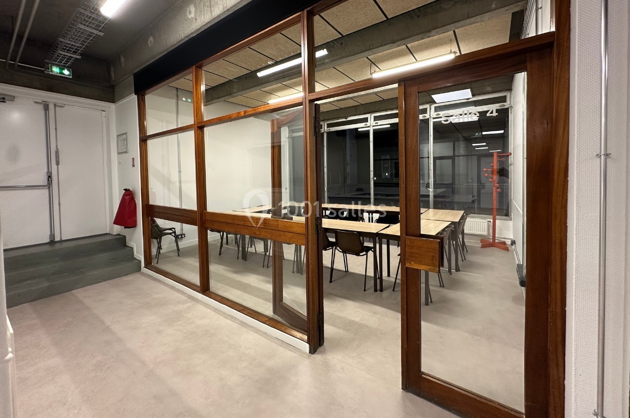 Salle de réunion vitrée avec tables, chaises et éclairage artificiel, vue depuis un couloir en béton.