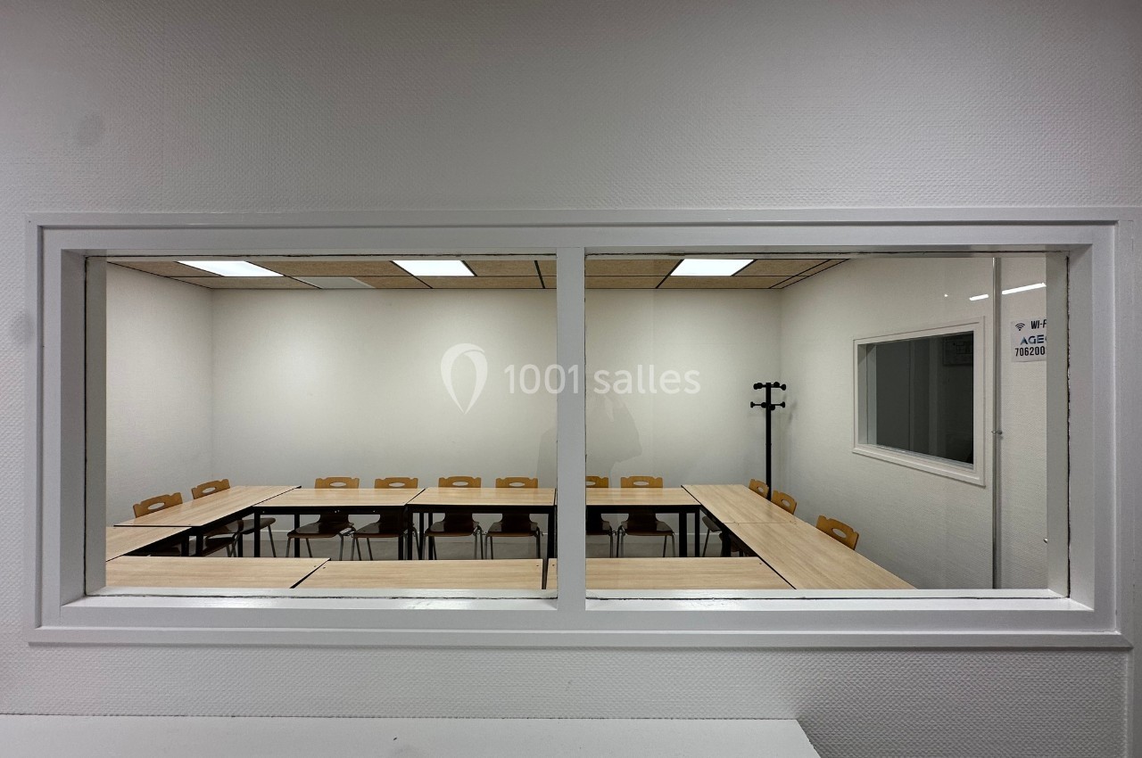 Salle de réunion vide avec des tables disposées en U, vue à travers une vitre dans un environnement sobre.