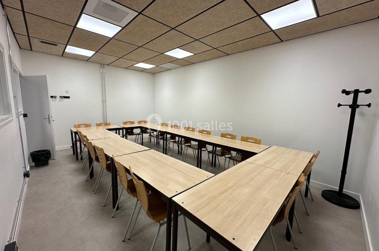 Salle de réunion avec tables disposées en U, chaises en bois, murs blancs et plafond avec dalles acoustiques.