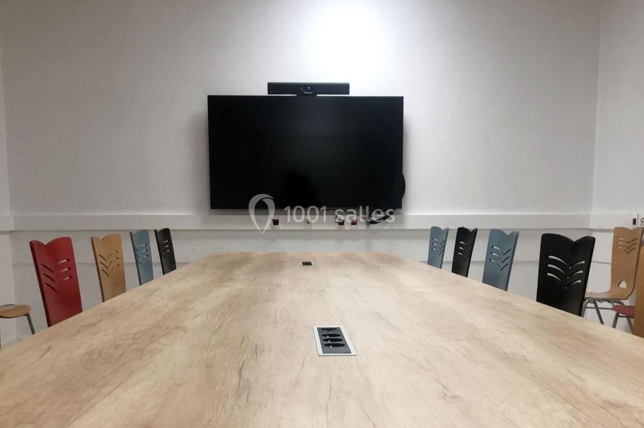 Salle de réunion avec une grande table en bois, des chaises colorées et un écran mural noir.