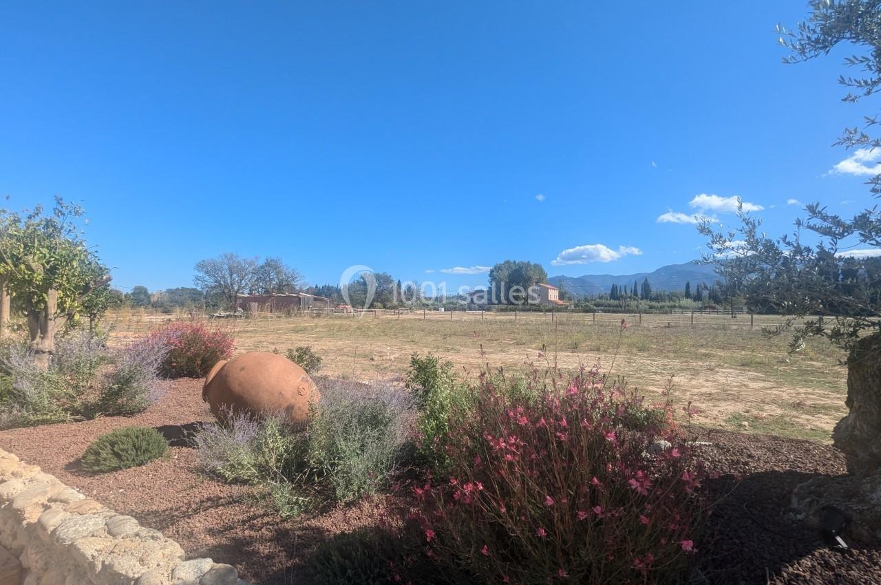 Jardin fleuri avec jarre en terre cuite au premier plan, vue dégagée sur un champ et des montagnes sous un ciel bleu.