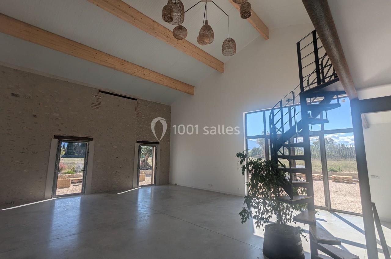 Intérieur lumineux avec plafond haut, poutres apparentes, escalier en colimaçon et grandes fenêtres donnant sur l'extérieur.