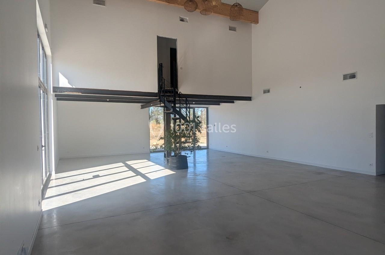 Intérieur d'une grande pièce lumineuse avec plafond haut, poutres apparentes, escalier central et sol en béton lisse.