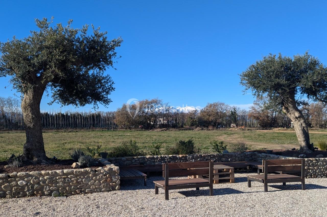 Deux bancs en bois sur un sol gravillonné, entourés d'arbres et d'un paysage champêtre sous un ciel bleu.