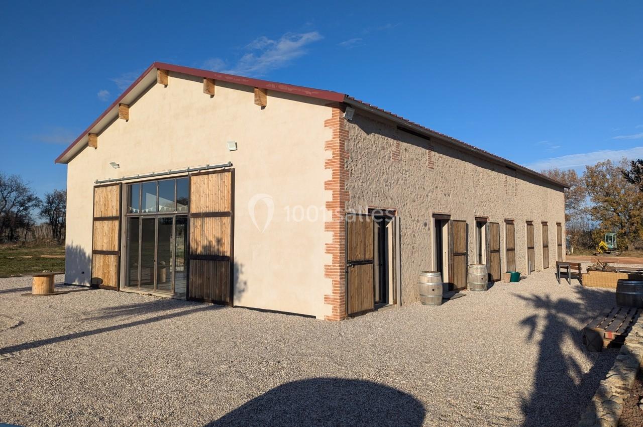 Bâtiment en pierre rénové avec des portes en bois, grandes baies vitrées et barils sur une cour en gravier.