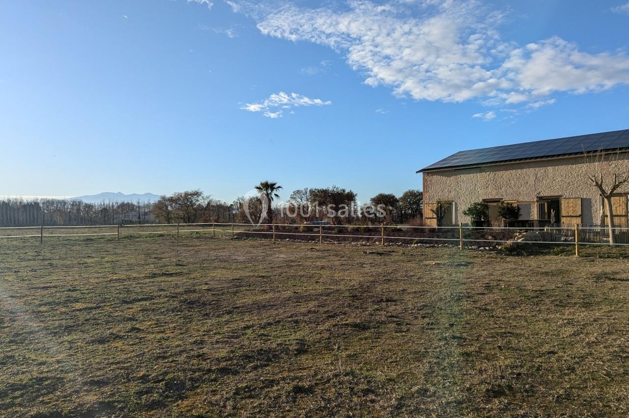 Champ herbeux avec une clôture, une maison en pierre à droite et un ciel bleu parsemé de nuages.