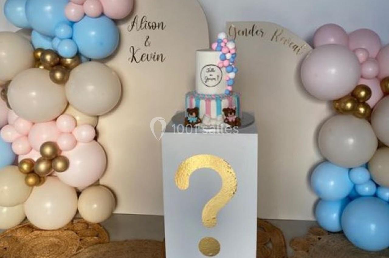 Décoration de fête avec ballons roses, bleus et dorés, gâteau sur piedestal blanc orné d'un point d'interrogation doré.