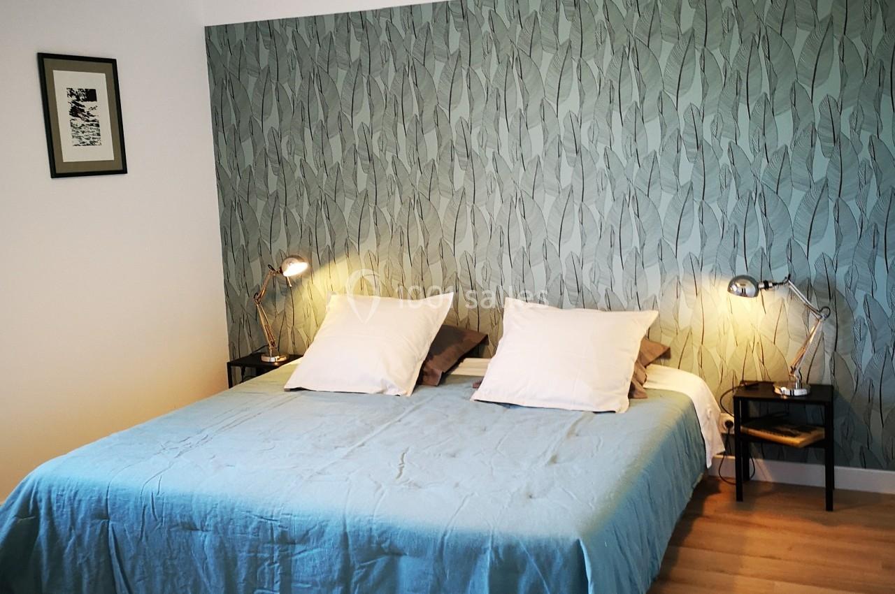 Lit double avec drap bleu, deux tables de chevet avec lampes et un mur décoré de papier peint à motifs végétaux.