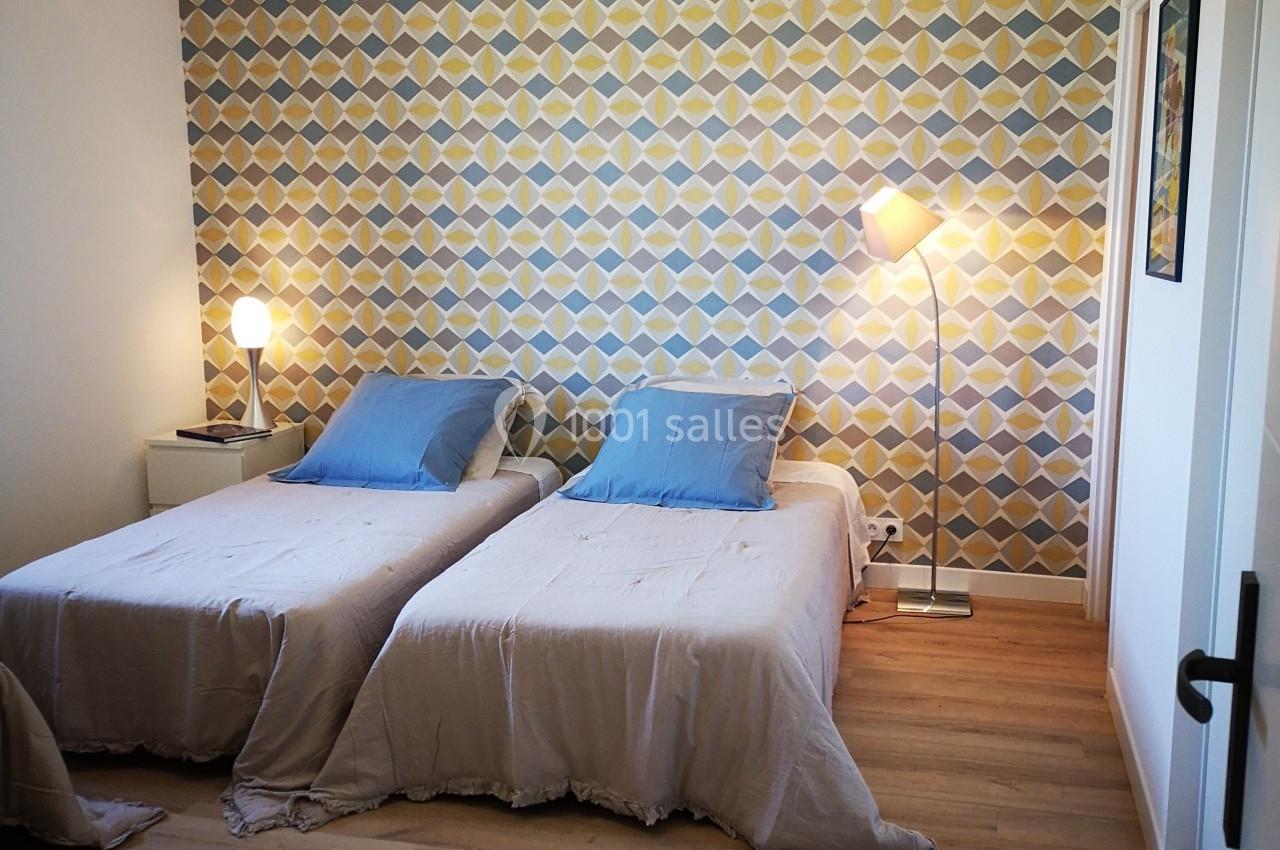 Deux lits simples avec draps beiges et oreillers bleus, éclairés par des lampes, devant un mur à motifs géométriques colorés.