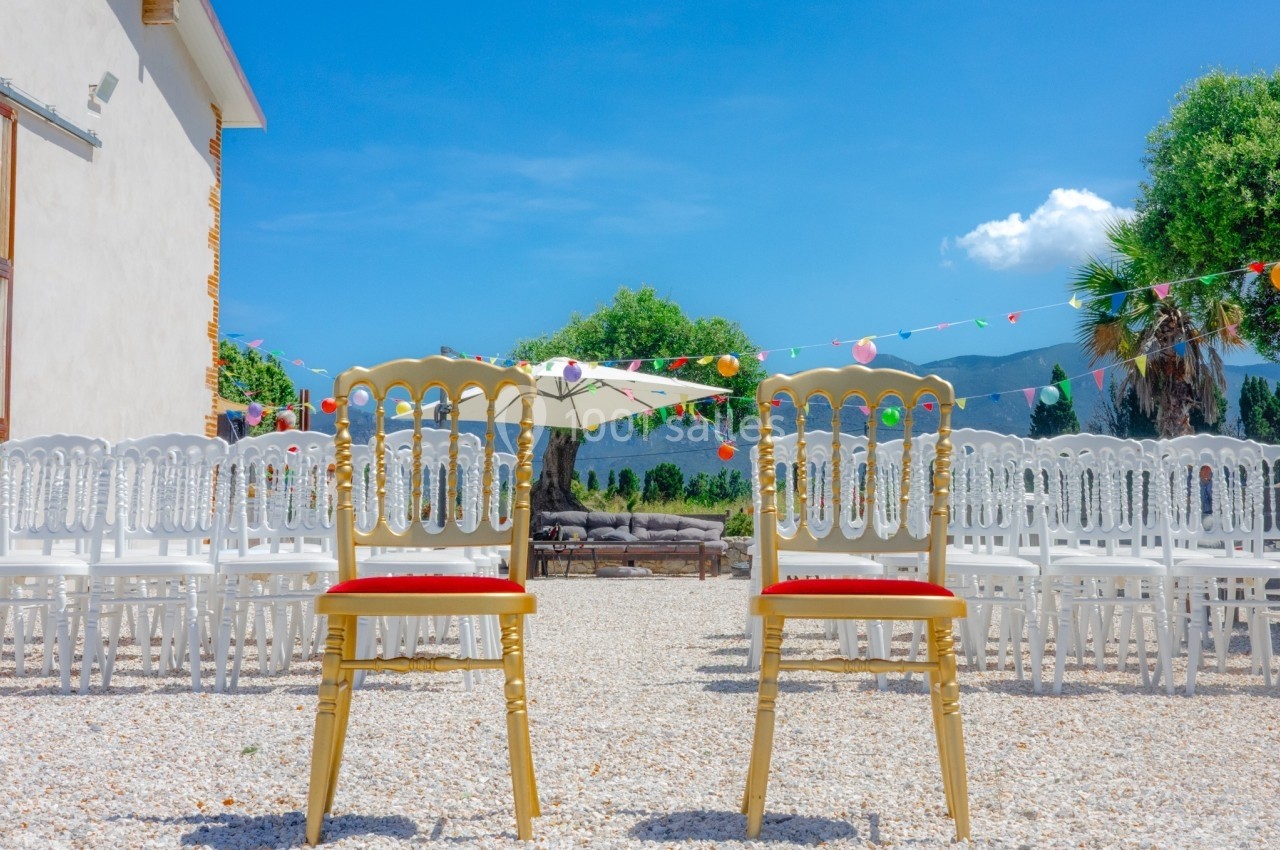 Deux chaises dorées alignées devant des rangées de chaises blanches en extérieur, sous un ciel bleu dégagé.