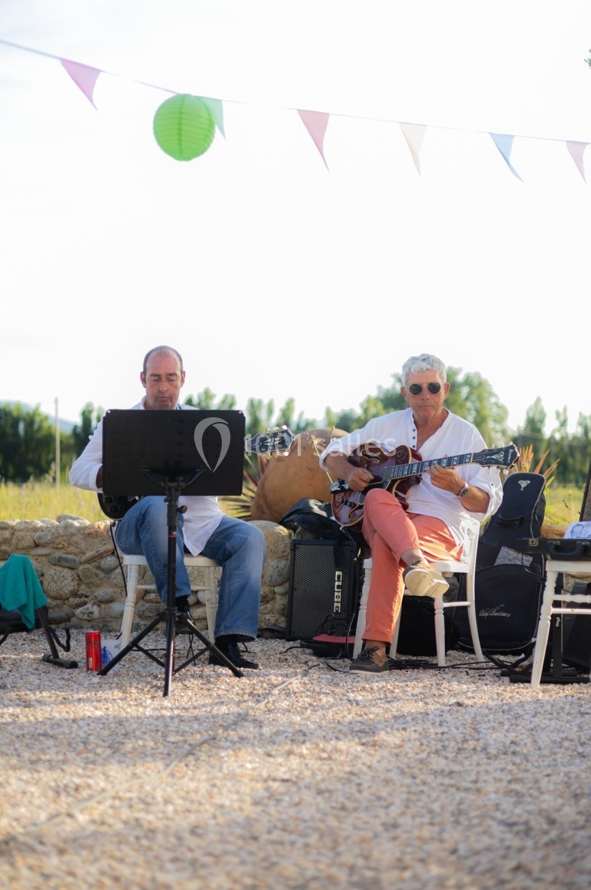 Deux musiciens jouent de la guitare en extérieur, assis devant un mur de pierre, sous des fanions colorés.