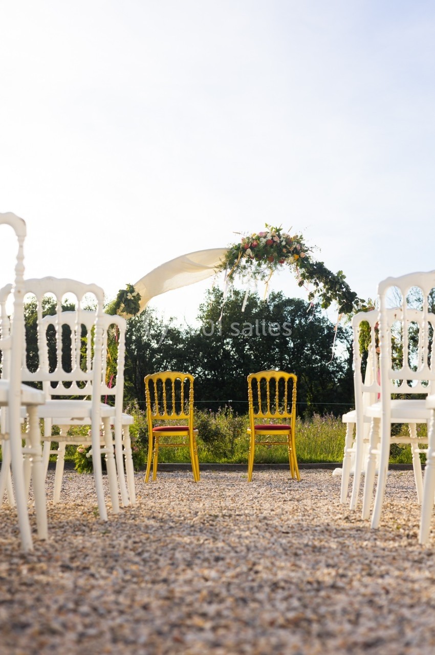 Deux chaises jaunes face à une arche fleurie, entourées de chaises blanches, dans un cadre extérieur ensoleillé.