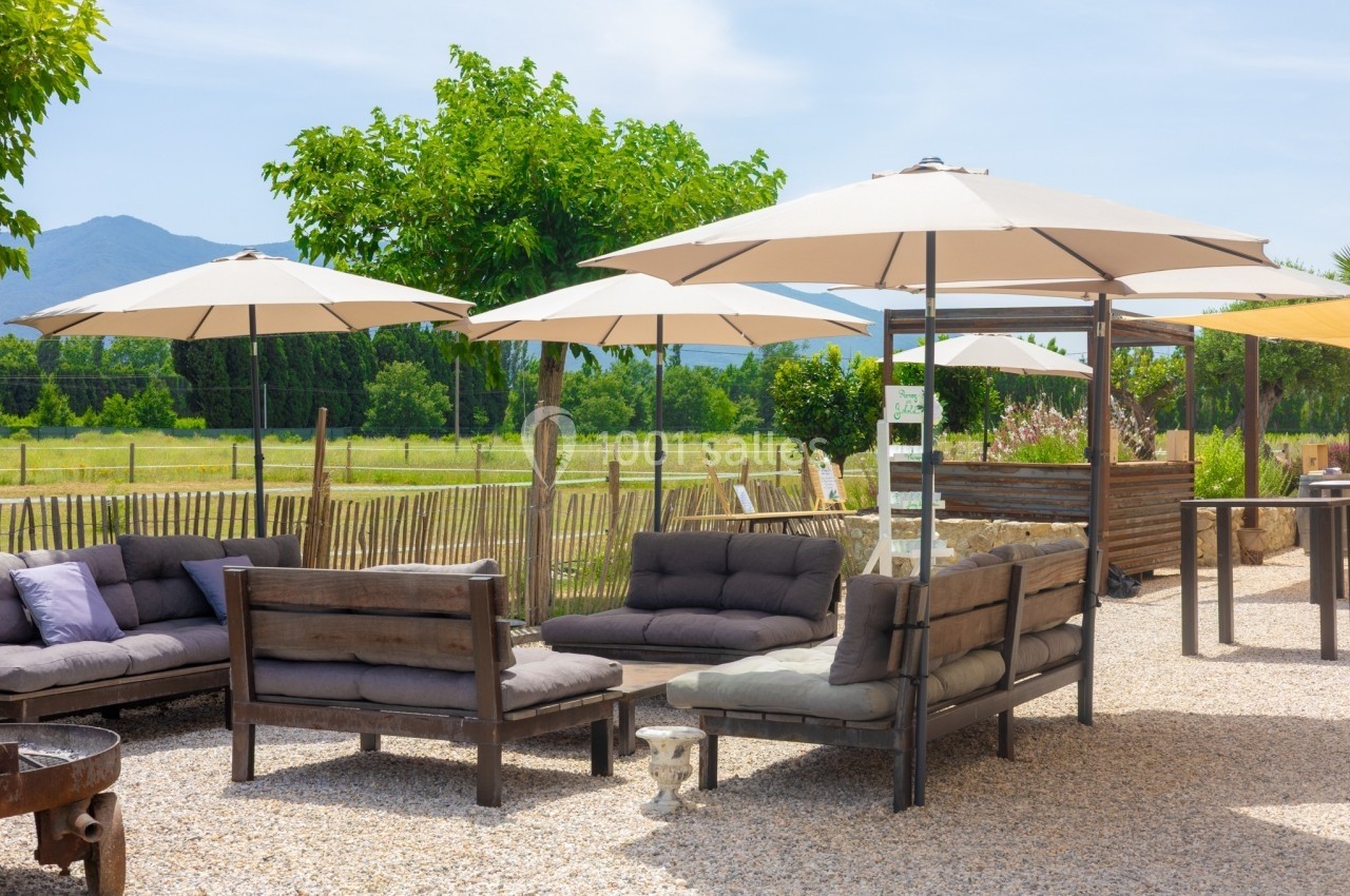 Mobilier de jardin avec canapés et parasols sur une terrasse en gravier, entourée de verdure et de champs.