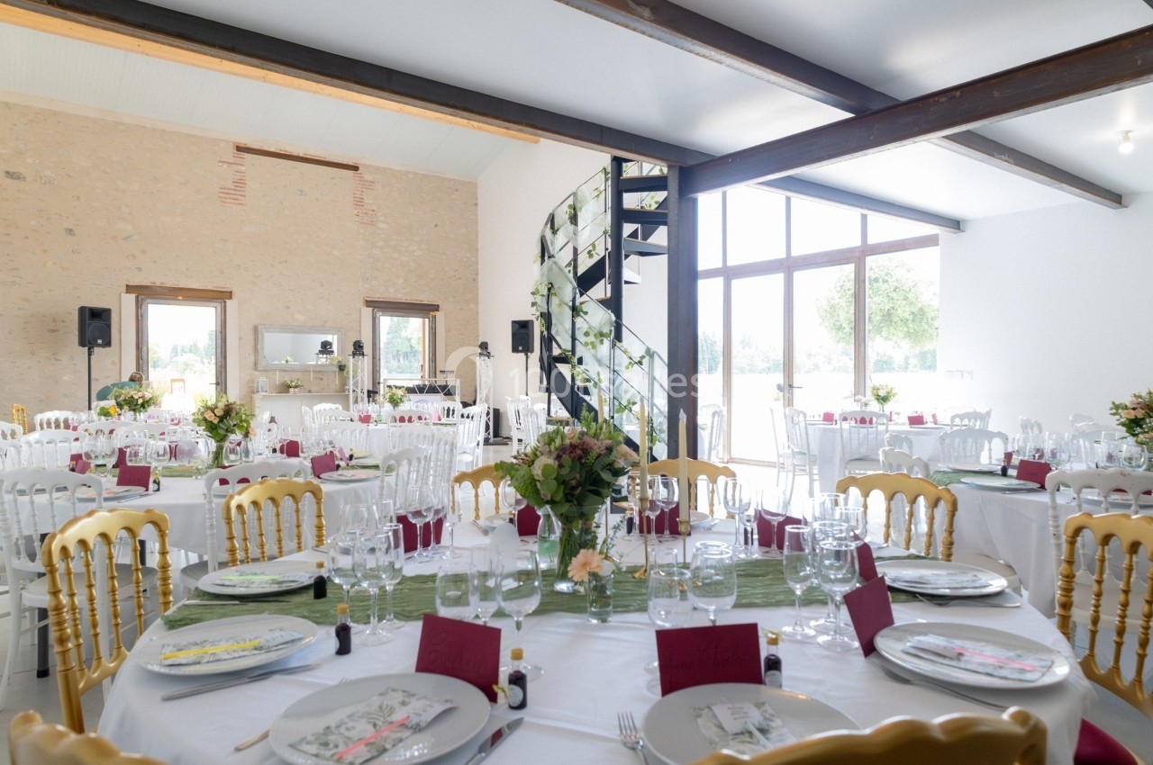 Salle de réception lumineuse décorée pour un mariage avec des tables rondes dressées et un escalier en colimaçon fleuri.