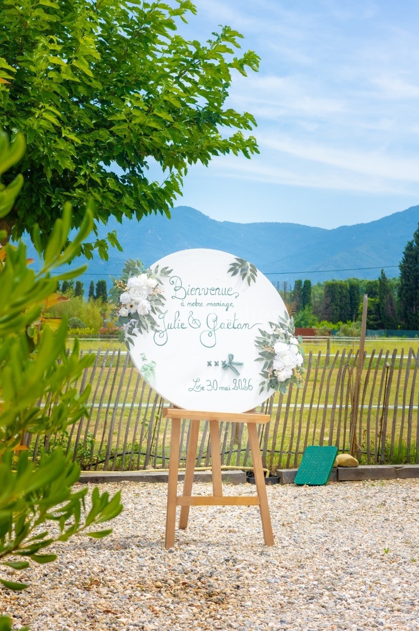 Panneau de bienvenue décoré de fleurs blanches sur un chevalet, dans un jardin avec vue sur des montagnes.