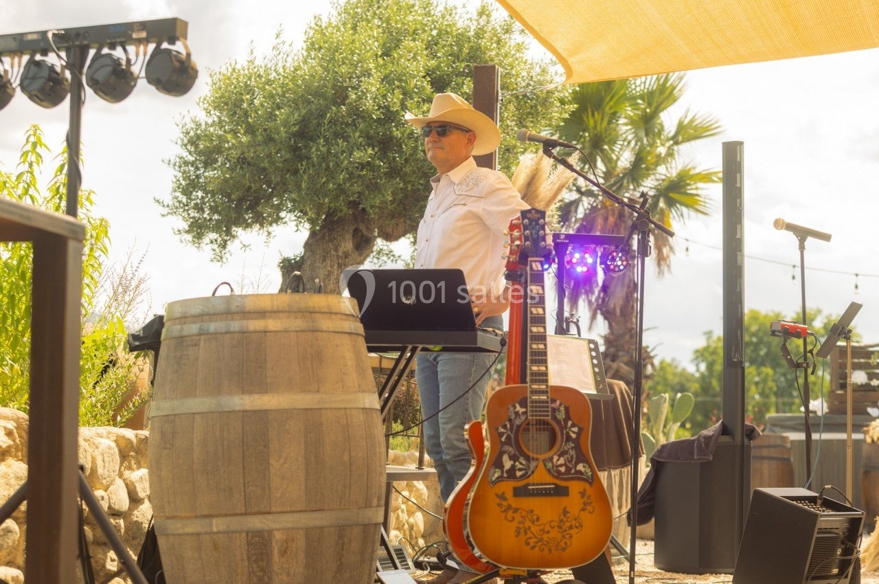 Un musicien avec un chapeau de cowboy joue du clavier en extérieur, près d'une guitare et d'un tonneau, sous un soleil…