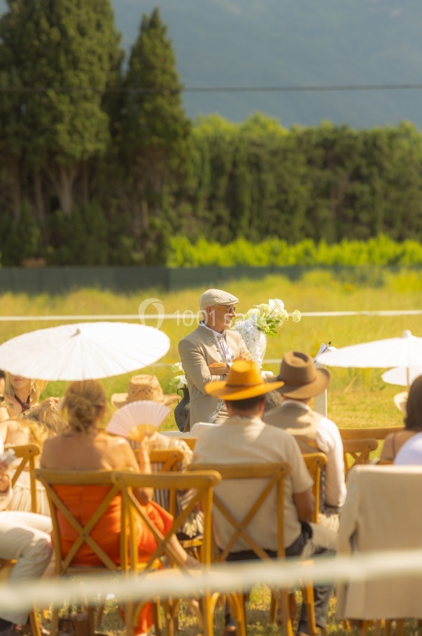 Un homme âgé s'exprime devant un public assis en plein air, sous un soleil estival, entouré de parasols et de verdure.