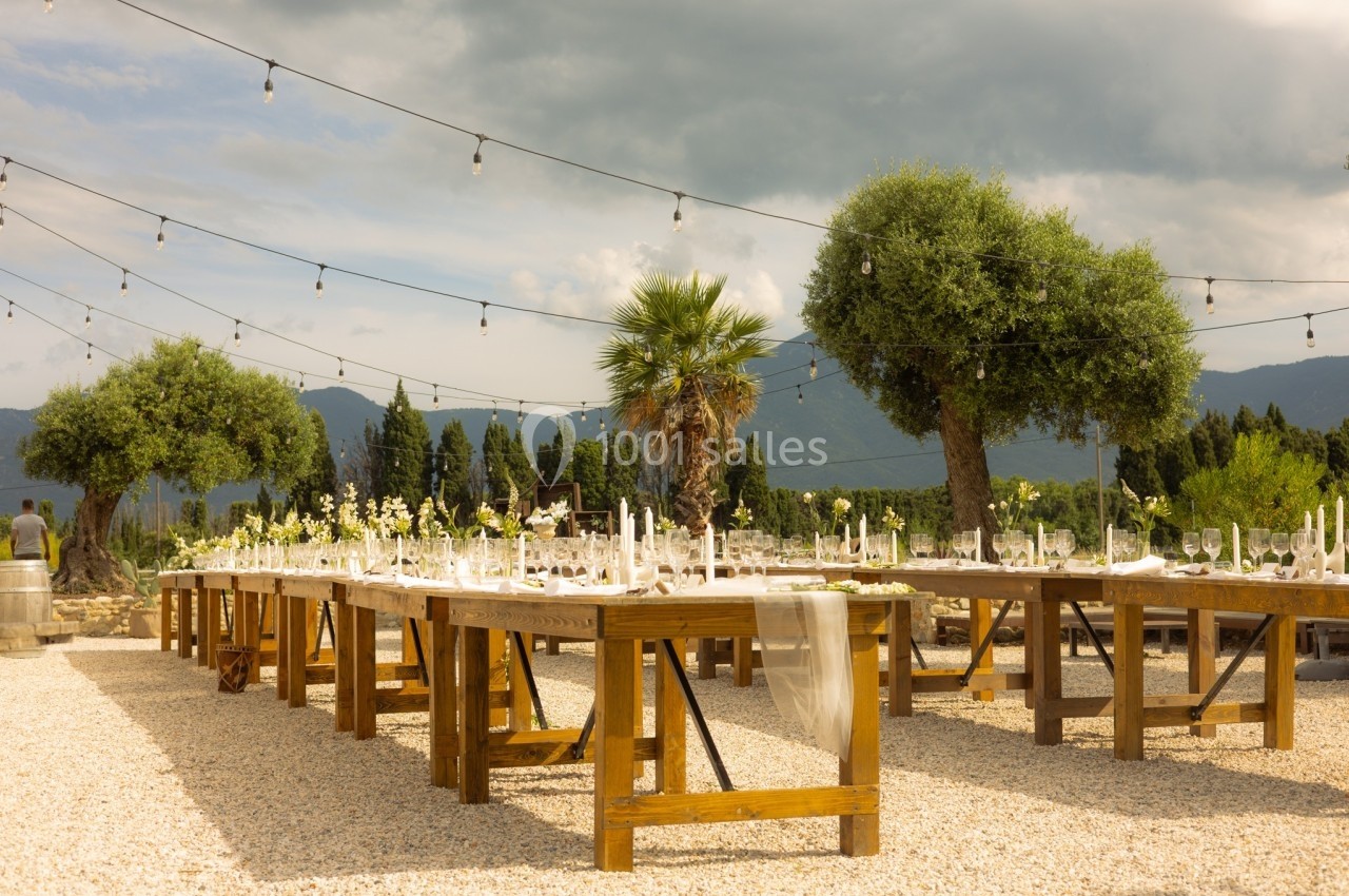 Tables en bois décorées pour un événement en extérieur, sous des guirlandes lumineuses, avec des arbres et des montagnes en…