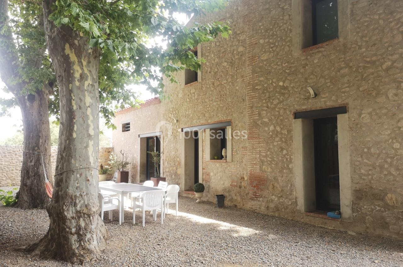 Cour ombragée avec des arbres, une table blanche entourée de chaises et une maison en pierre aux grandes fenêtres.