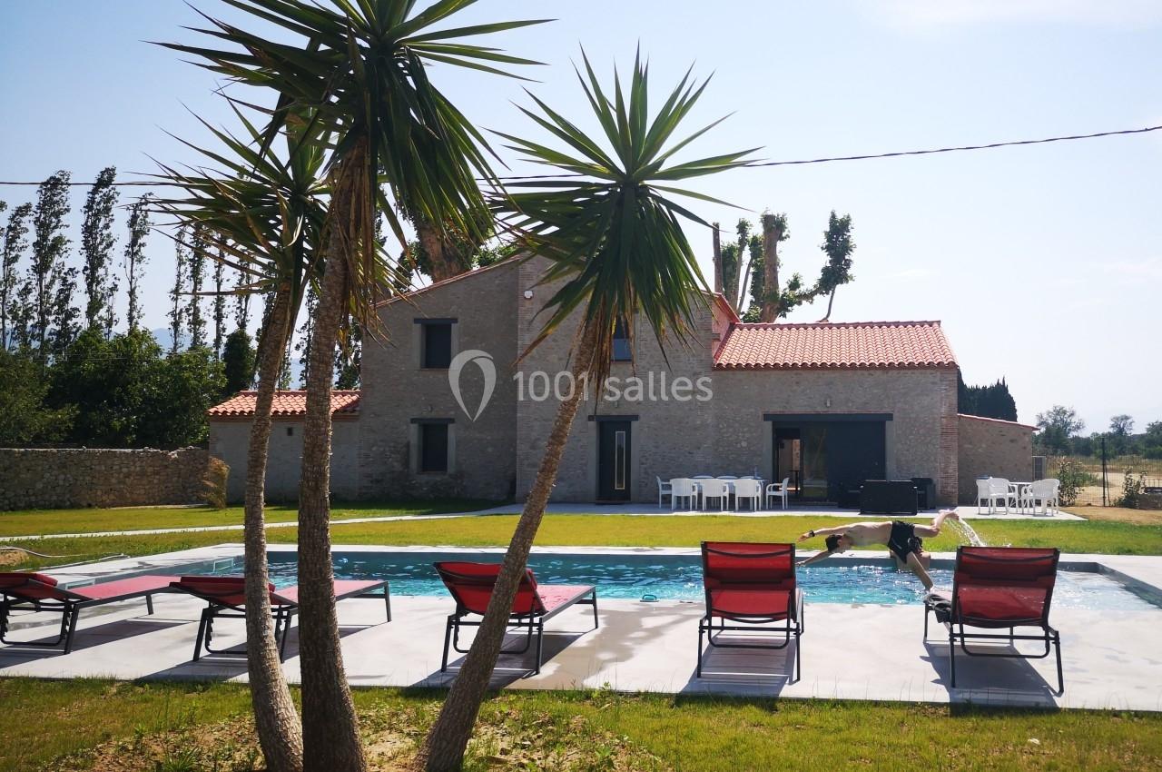 Maison en pierre avec piscine, transats rouges et palmiers au premier plan, située dans un cadre rural ensoleillé.