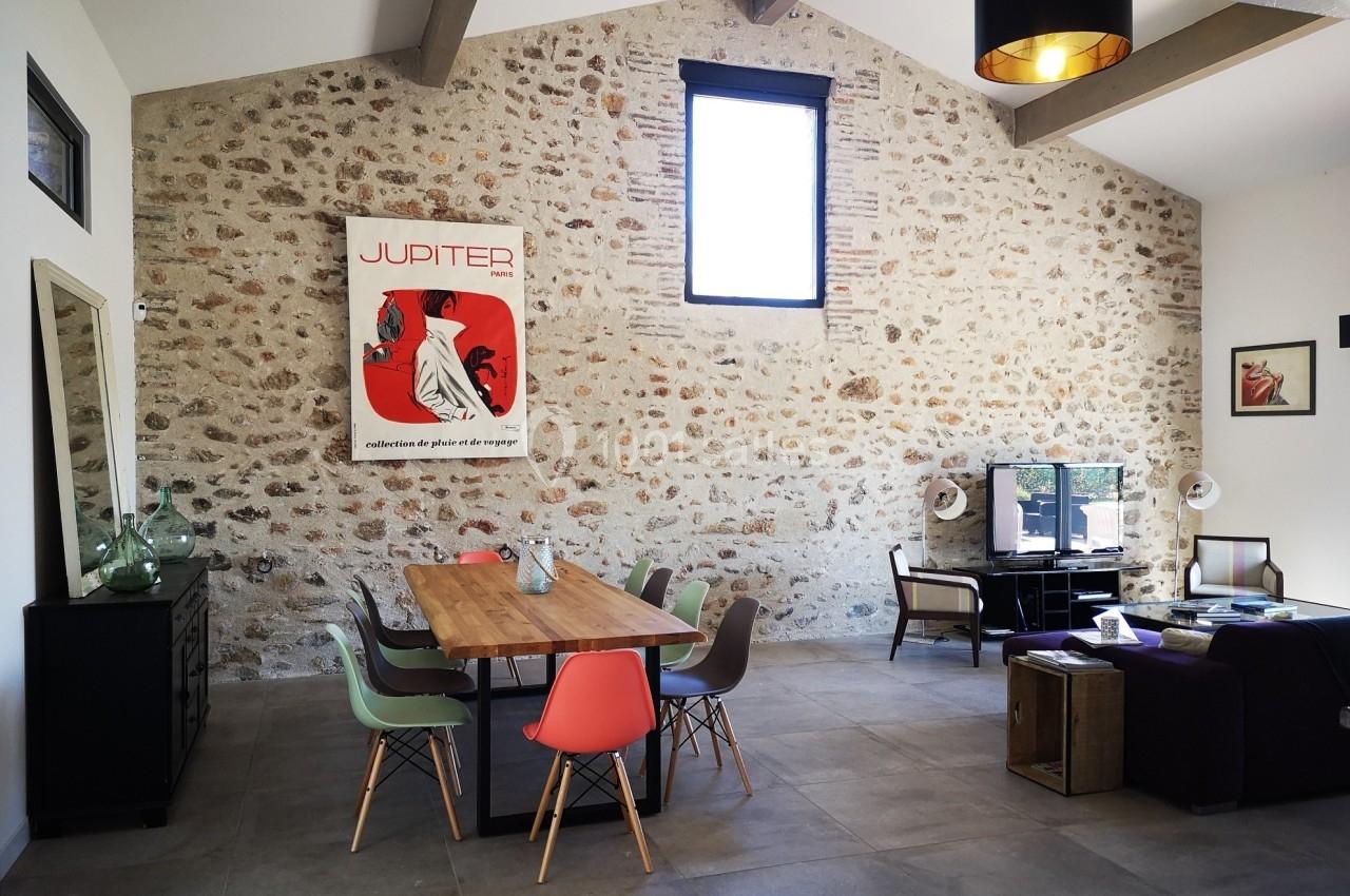 Salle à manger lumineuse avec table en bois, chaises colorées, mur en pierre apparente et coin salon avec télévision.