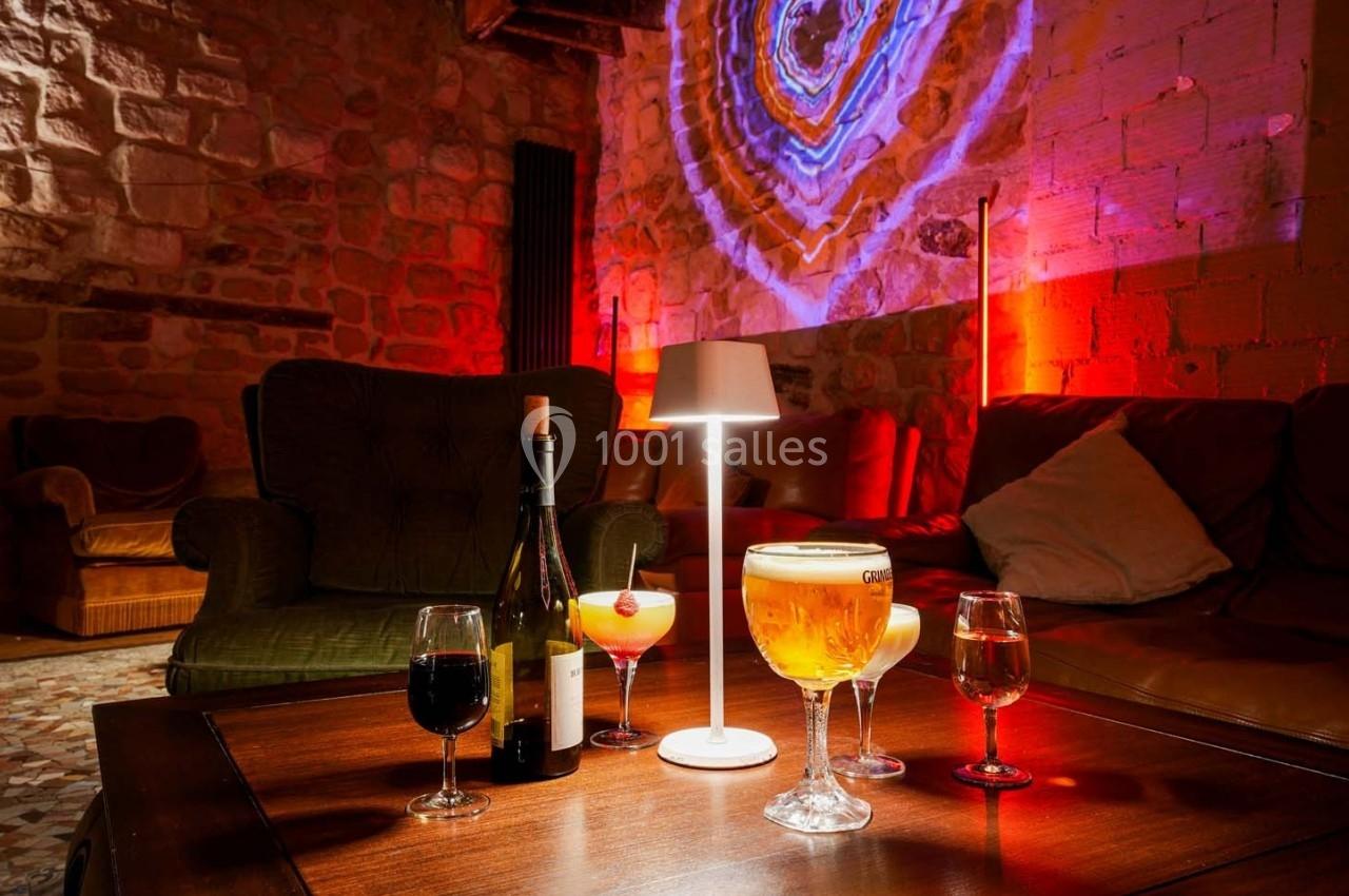 Table basse avec divers verres de boissons, une bouteille de vin et une lampe, dans un salon à l'éclairage tamisé.