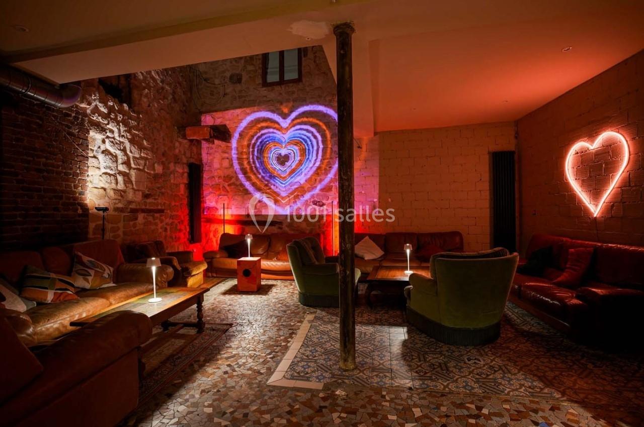 Salle cosy avec murs en briques, fauteuils et canapés, éclairée par des néons en forme de cœur et des lampes d'appoint.