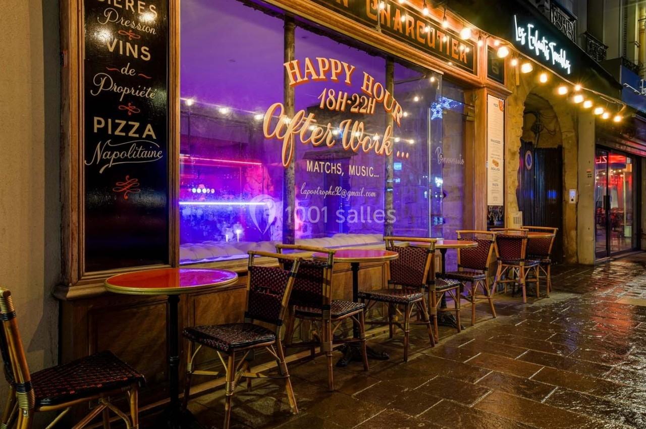 Façade d'un café-restaurant avec terrasse vide, éclairée par des lumières colorées en soirée pluvieuse.