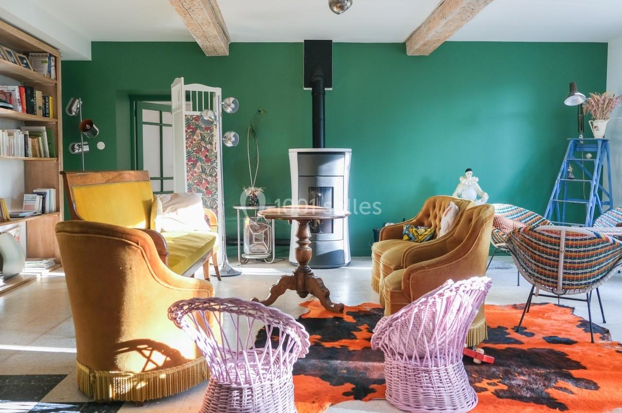 Salon coloré avec murs verts, mobilier vintage, poêle à bois central, tapis orange et noir, et étagères remplies de livres.