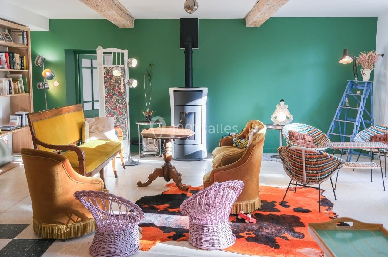 Salon lumineux avec murs verts, poêle à bois, mobilier vintage coloré et tapis orange sur sol clair.
