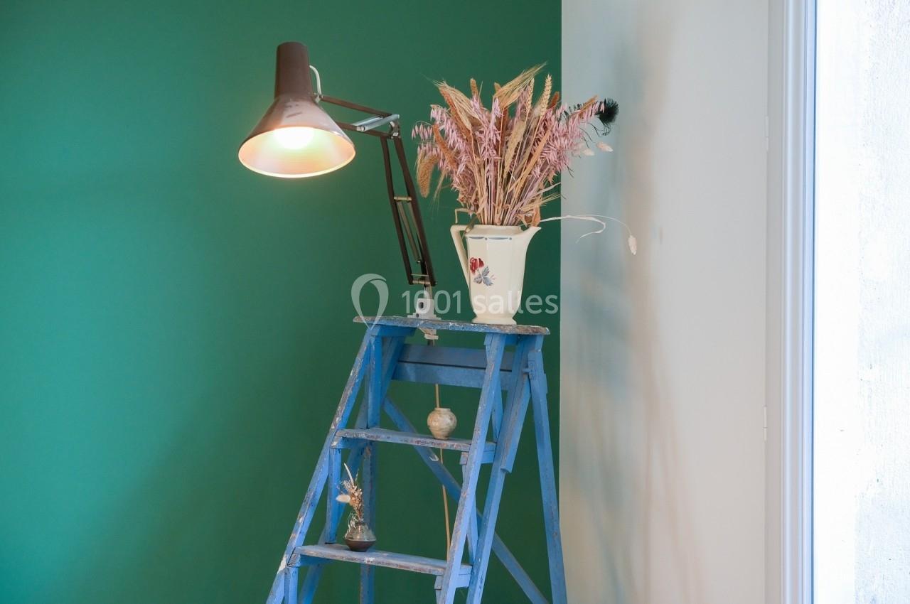 Lampe allumée sur une échelle bleue avec un vase de fleurs séchées, devant un mur vert et blanc.