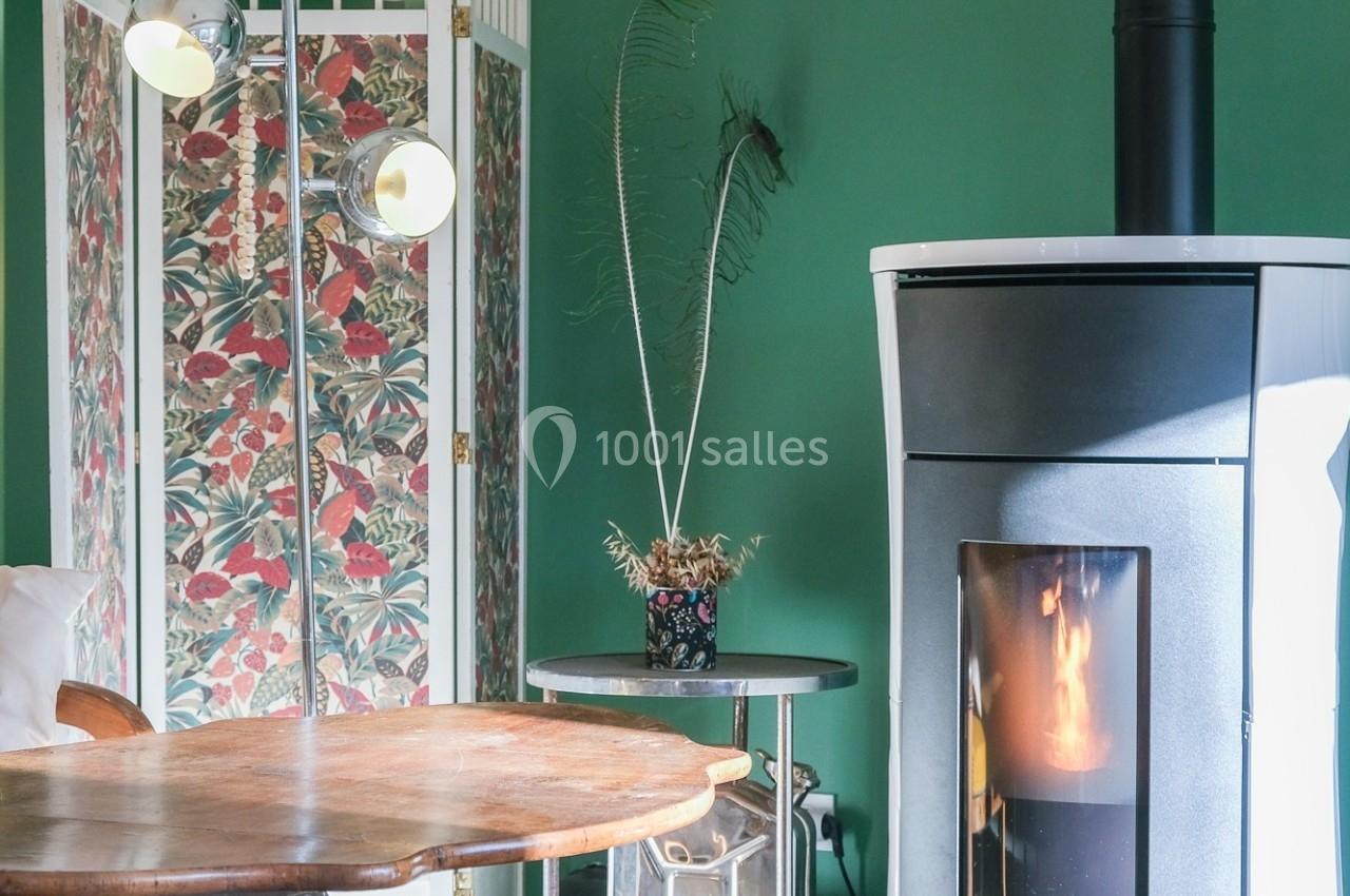 Poêle à bois allumé dans un salon avec mur vert, paravent fleuri, table en bois et fauteuils.