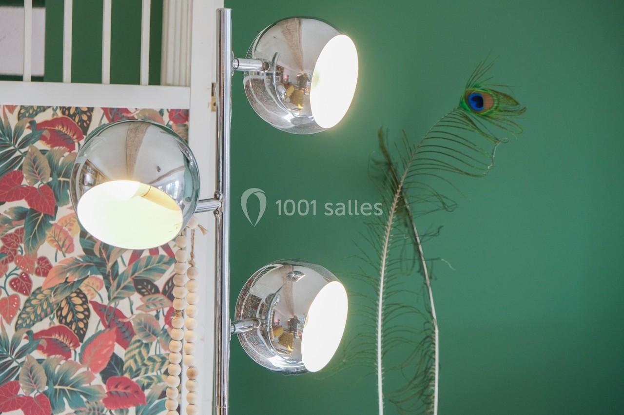 Lampadaire chromé à trois globes lumineux devant un mur vert, décoré d'un motif floral et d'une plume de paon.