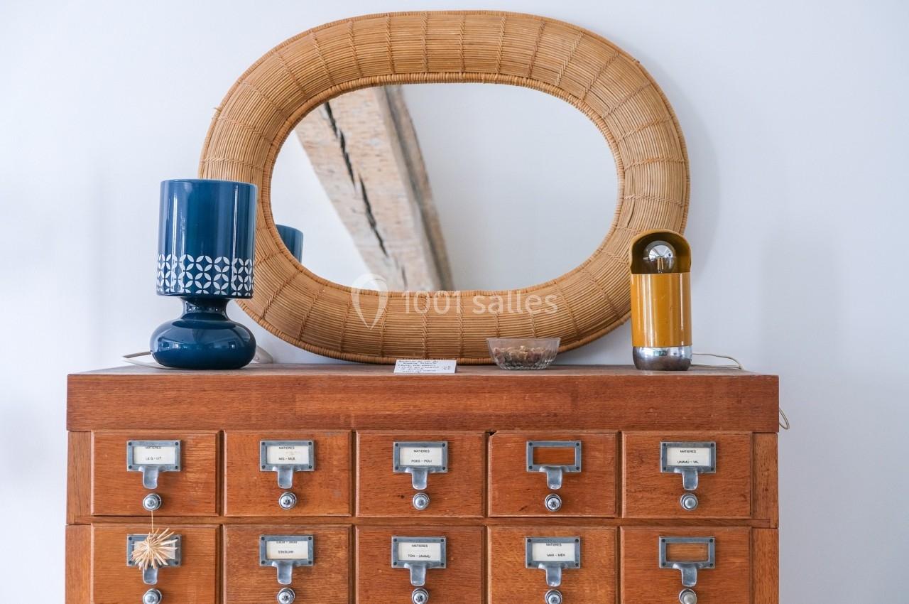 Meuble en bois à tiroirs étiquetés, surmonté d'un miroir ovale en rotin, d'une lampe bleue et d'une lampe jaune.