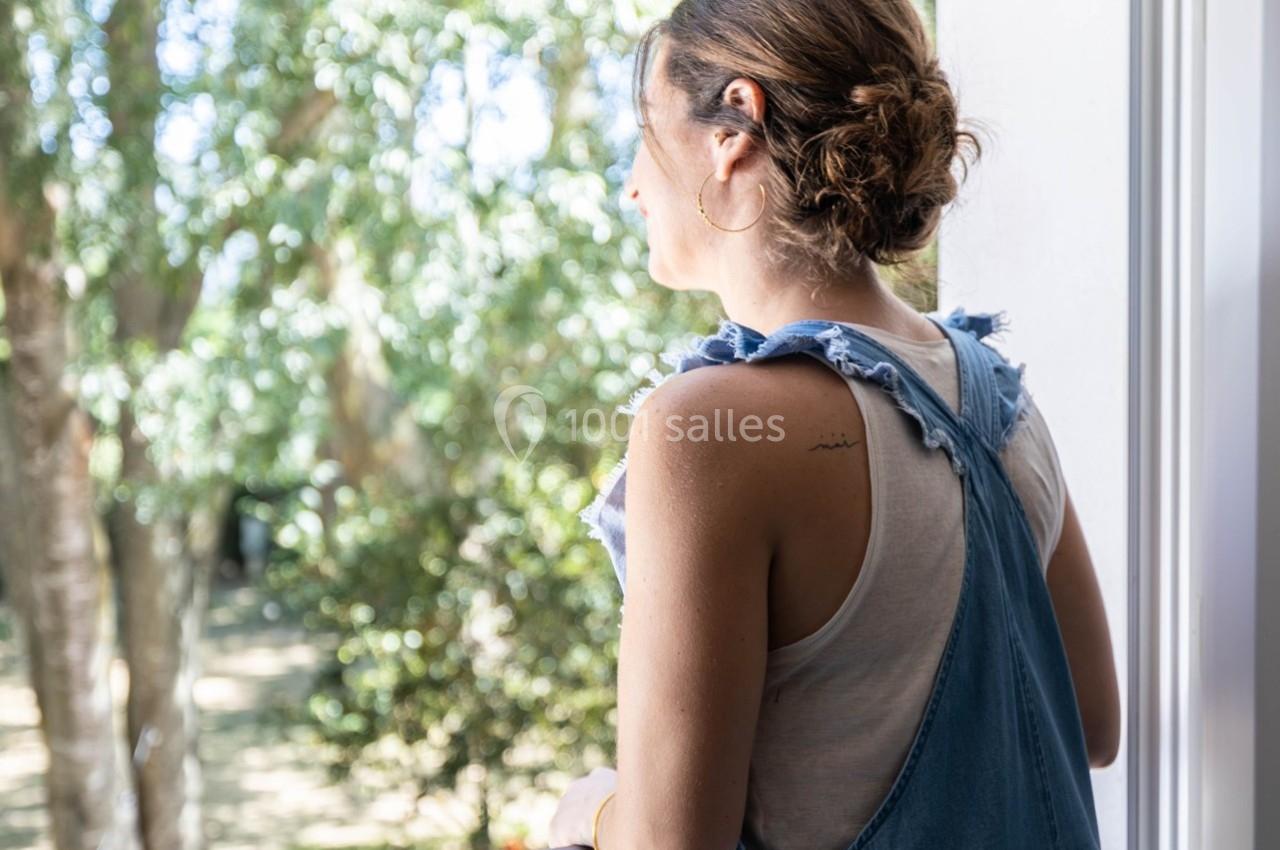 Femme de dos regardant un jardin verdoyant depuis un balcon, portant une salopette en jean et un haut blanc.