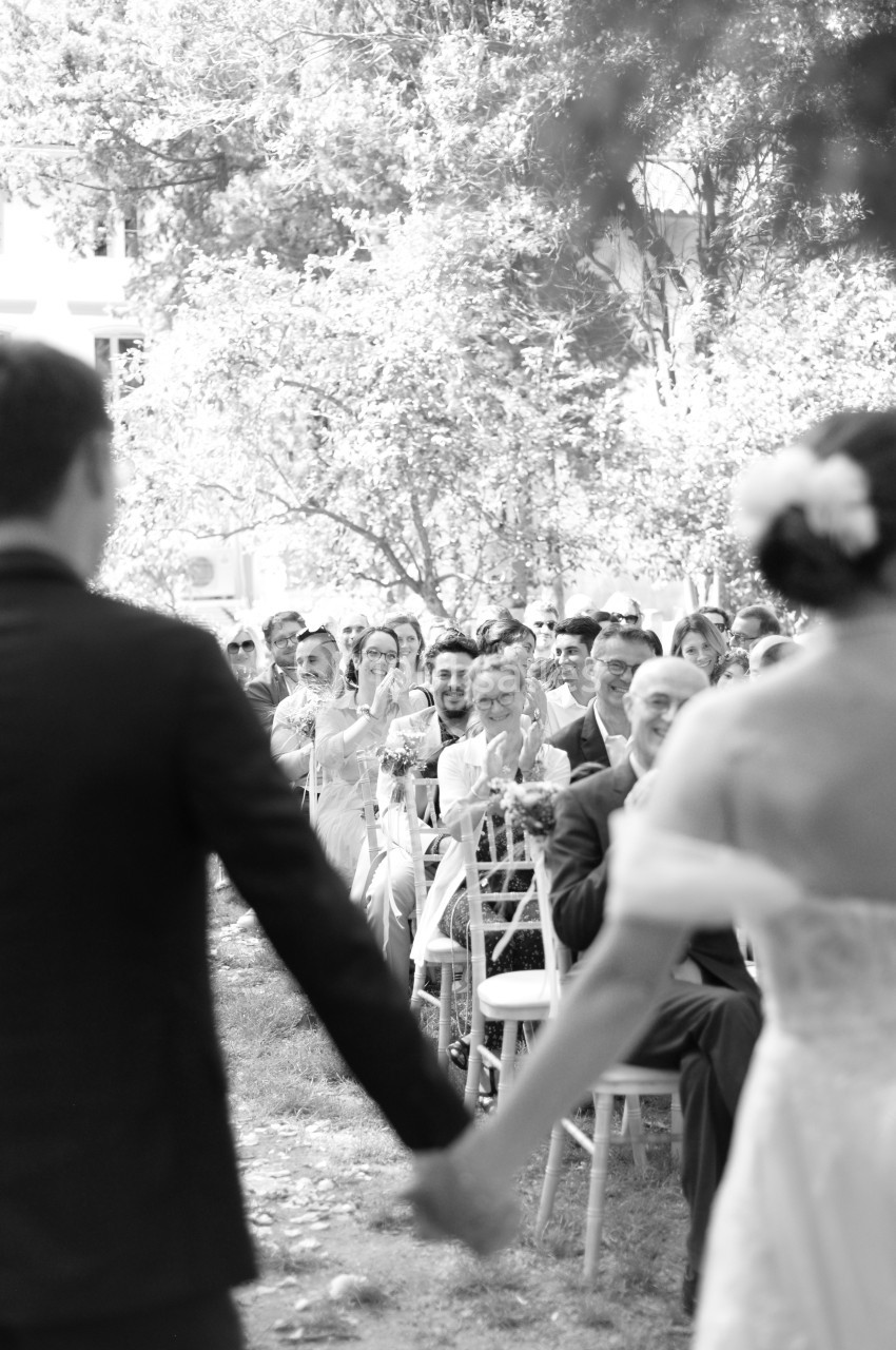 Un couple de mariés avance main dans la main vers des invités assis à l'extérieur lors d'une cérémonie.