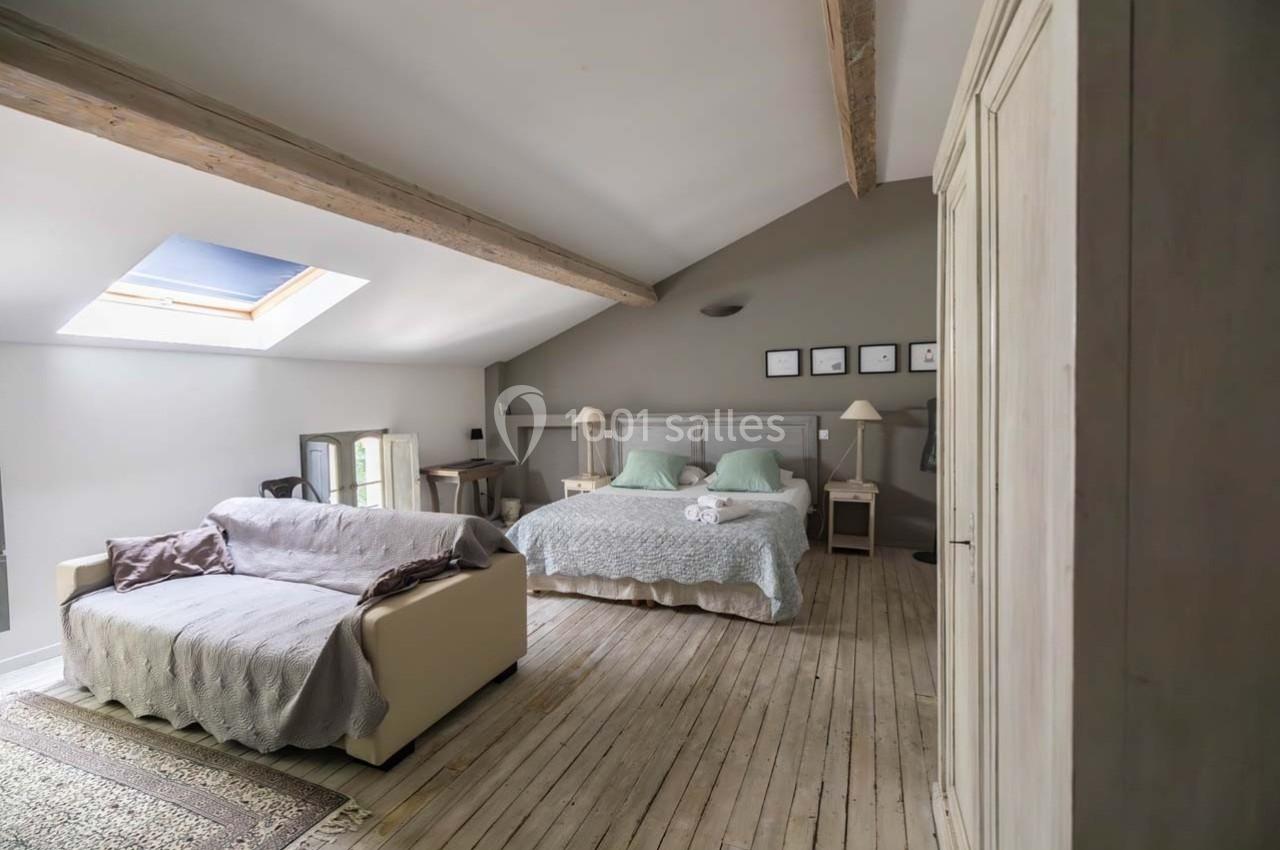 Chambre lumineuse avec lit double, canapé beige, poutres apparentes et parquet clair.