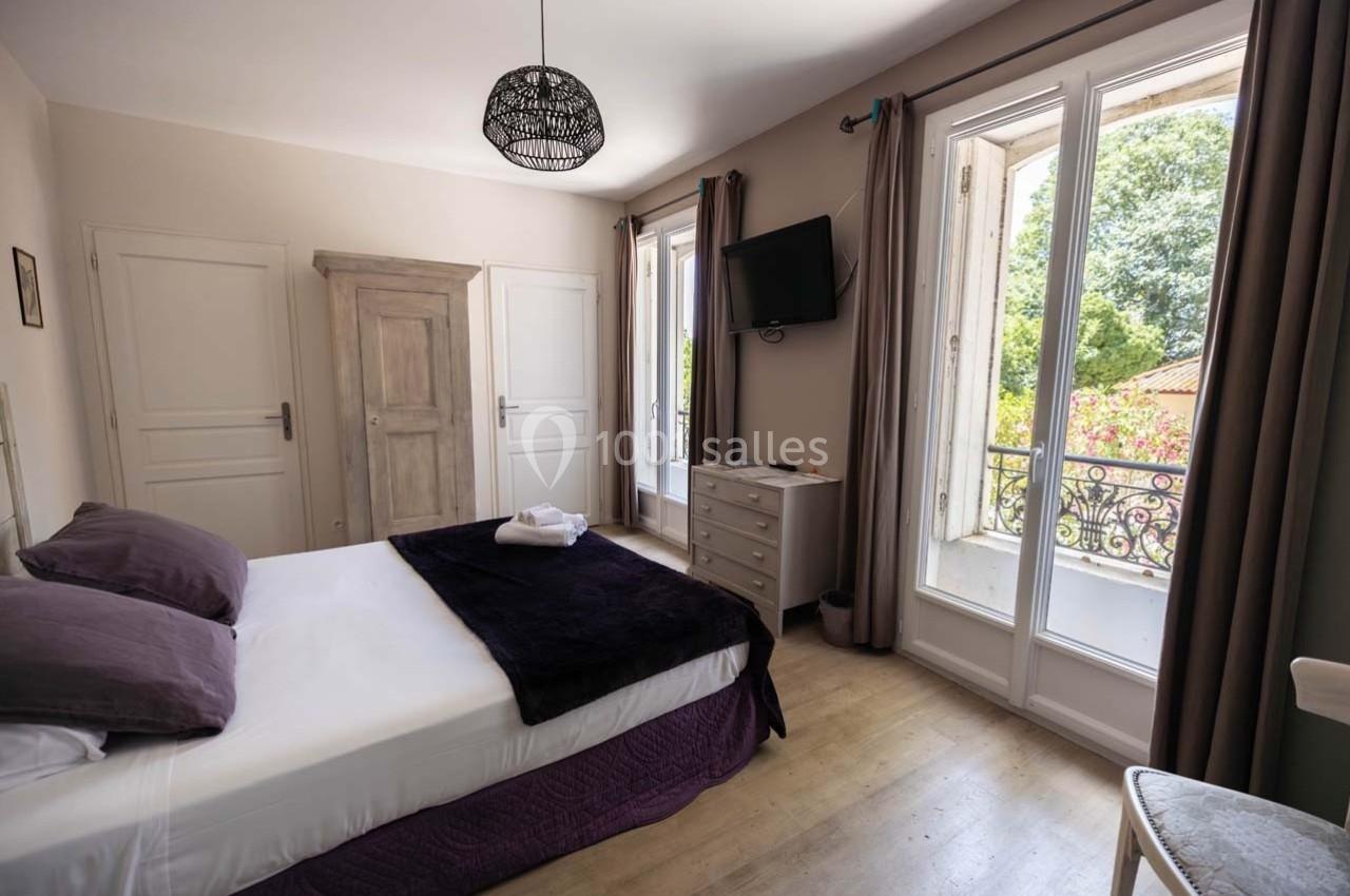 Chambre lumineuse avec lit double, armoire en bois, télévision murale et fenêtres donnant sur un balcon avec vue sur jardin.