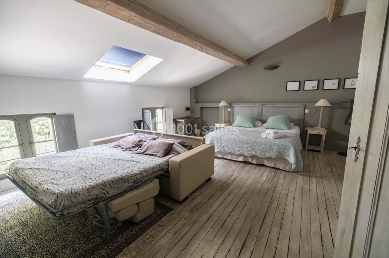 Chambre lumineuse avec parquet, un canapé-lit déplié et un grand lit double, sous un plafond avec poutres apparentes.