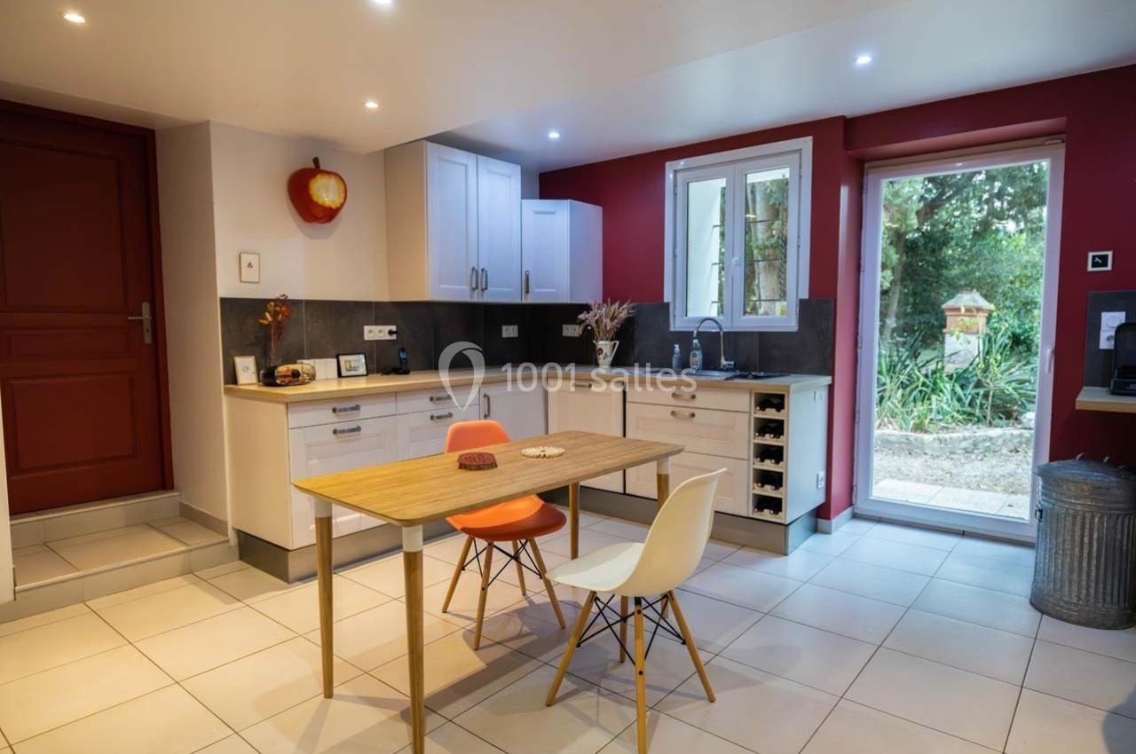 Cuisine moderne avec meubles blancs, table en bois, deux chaises, mur rouge, et vue sur un jardin par une porte vitrée.