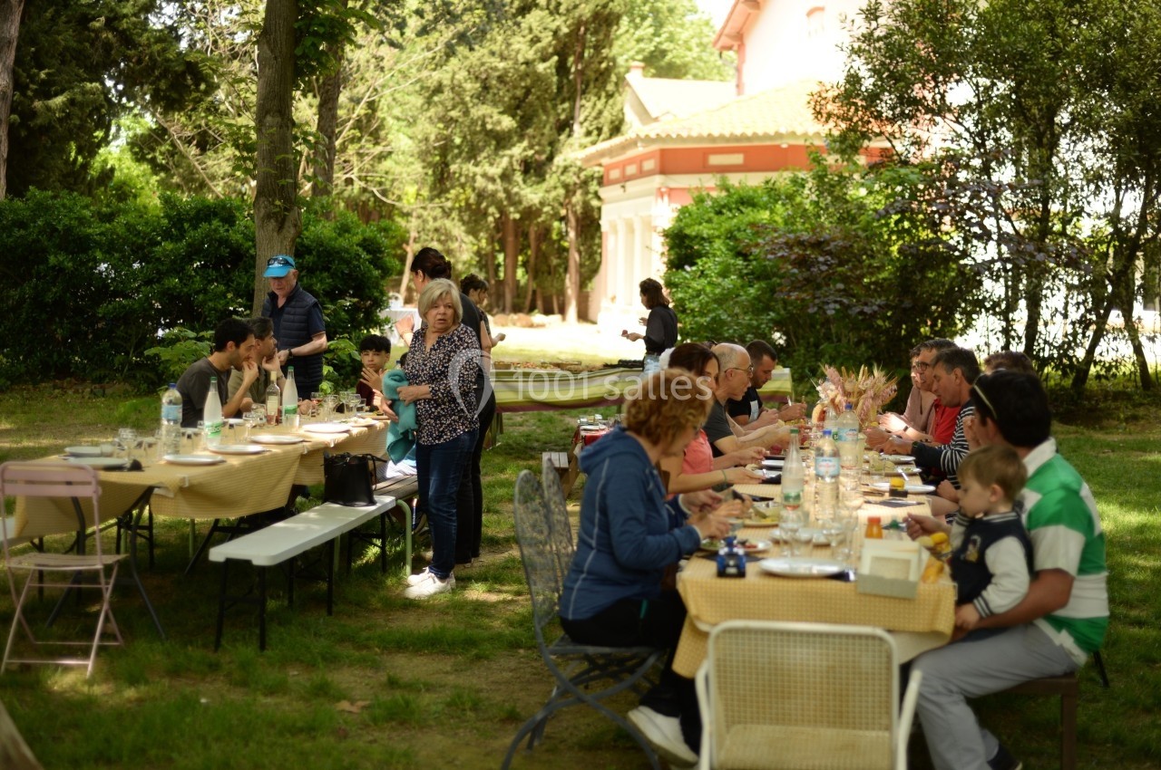 Des personnes partagent un repas en plein air autour de longues tables dans un jardin arboré.