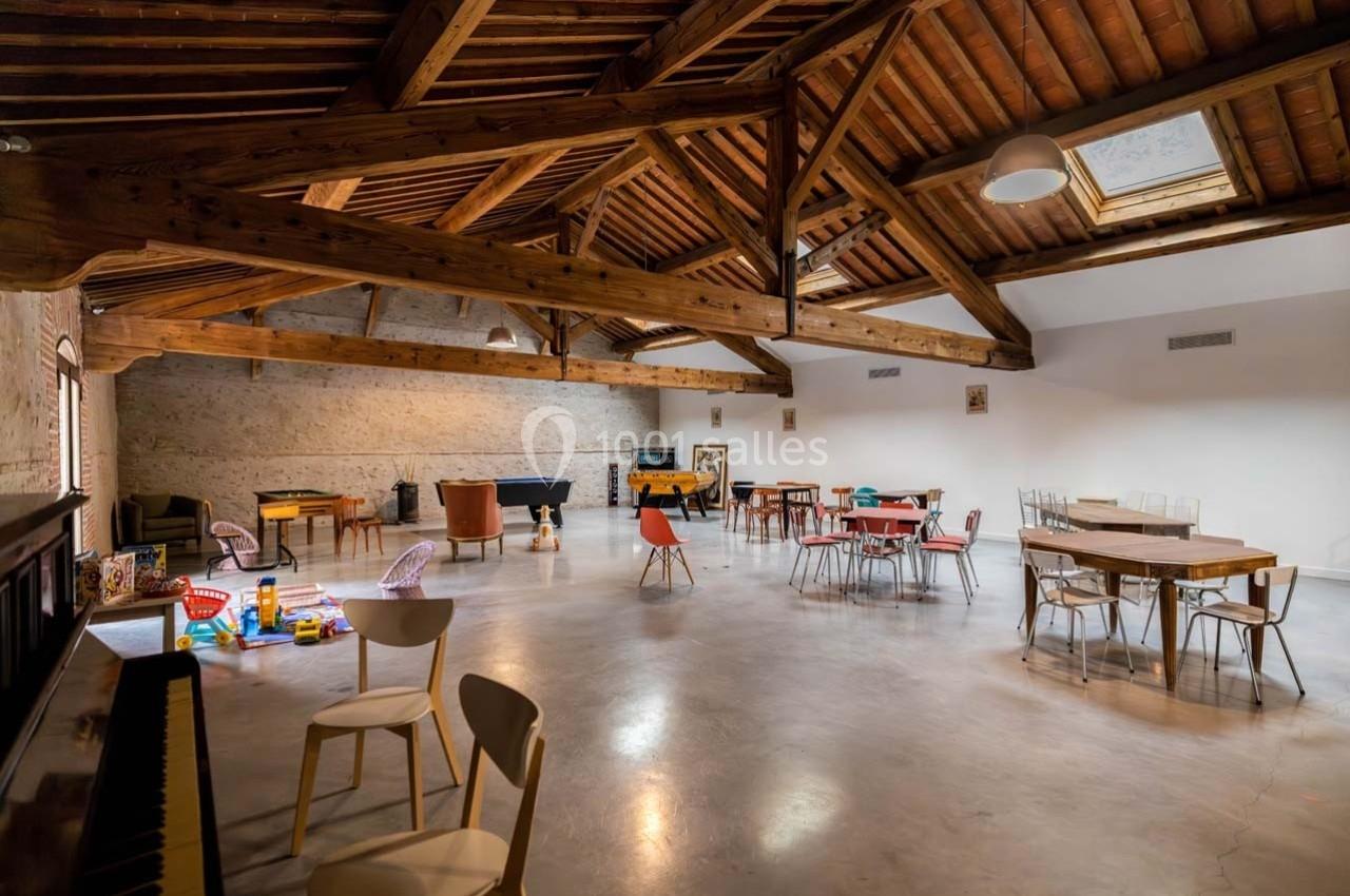 Salle spacieuse avec poutres apparentes, tables, chaises, jeux pour enfants et lumière naturelle provenant d'une lucarne.