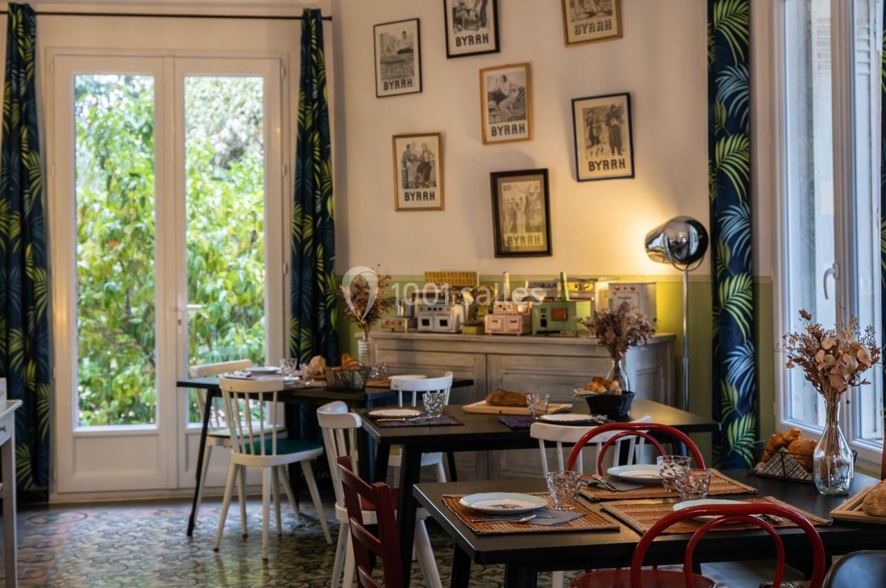 Salle à manger lumineuse avec tables dressées, chaises colorées, décor rétro et vue sur un jardin verdoyant.