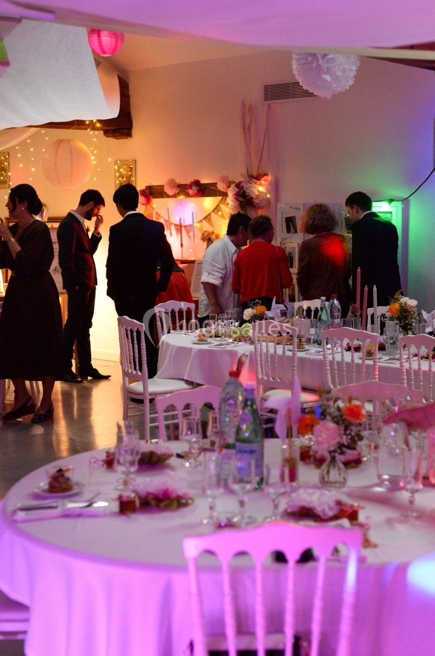 Salle décorée pour un événement avec des tables dressées et des invités debout discutant près d'un buffet.