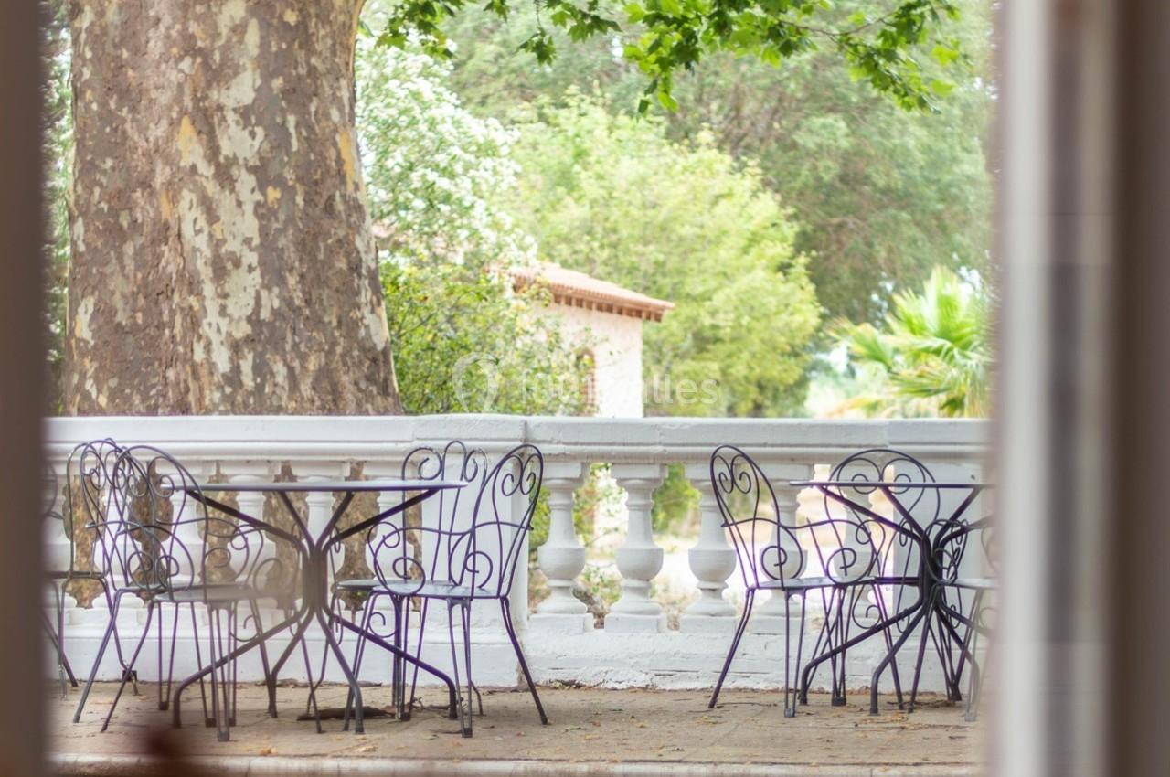 Terrasse avec tables et chaises en fer forgé, entourée de verdure et bordée d'une balustrade blanche.