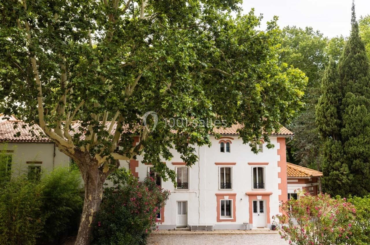Façade d'une maison blanche avec des volets et encadrements rouges, entourée d'arbres et de végétation.