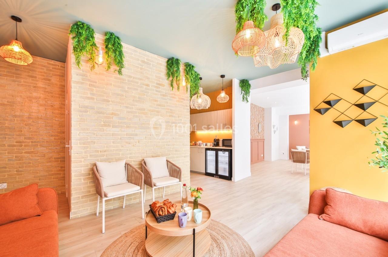 Salon lumineux avec murs en briques et jaunes, mobilier moderne, plantes suspendues et vue sur une cuisine ouverte.